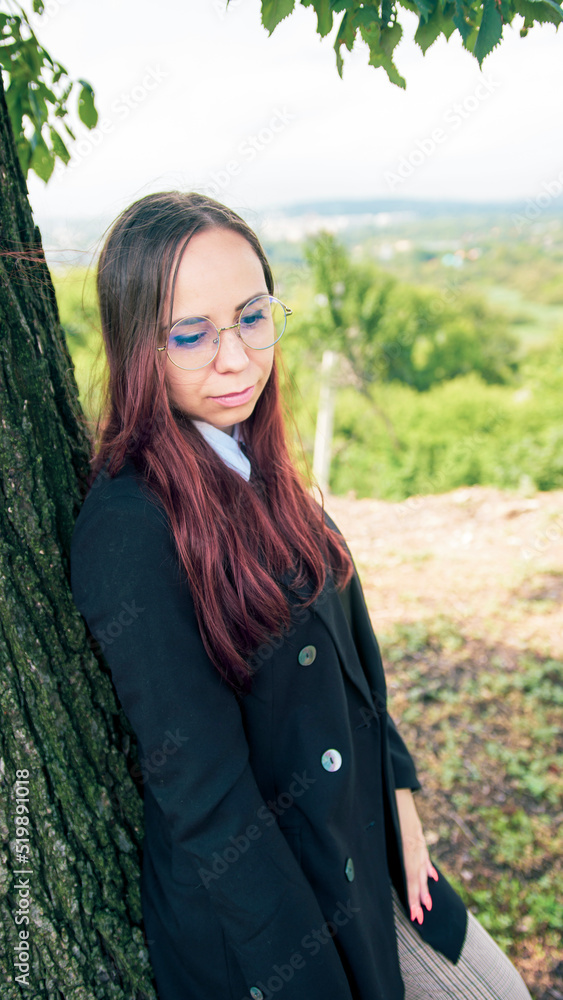 Young woman in glasses, business suit standing near tree in nature. Business woman posing near tree trunk during break on background of countryside.
