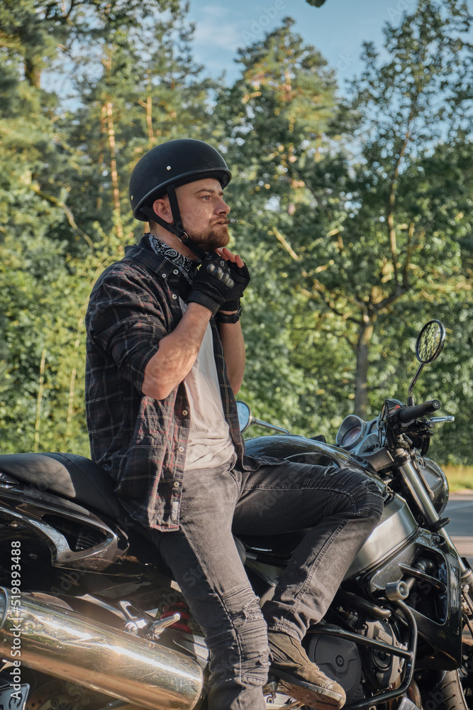 A young male biker travels on a motorcycle alone, stopped and puts on a helmet on the side of a forest road A young male biker travels on a motorcycle alone, stopped and puts on a helmet on the side of a forest road