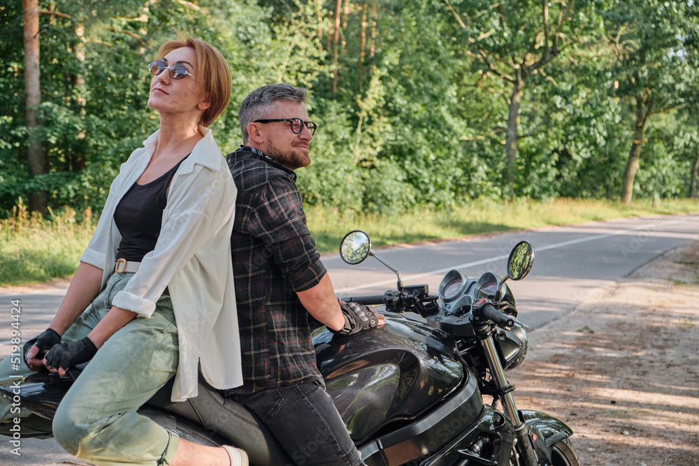 Middle age couple talking and having fun, sitting on a motorcycle, traveling together on a forest road Middle age couple talking and having fun, sitting on a motorcycle, traveling together on a forest road