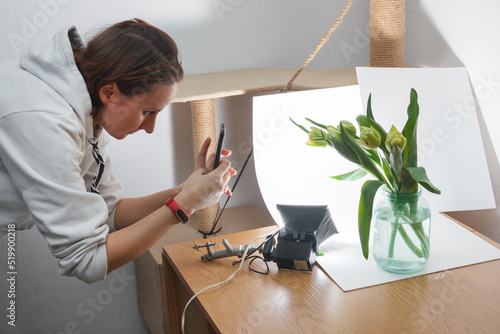 The young girl takes pictures of flowers in the improvised home photo studio using the smartphone.
