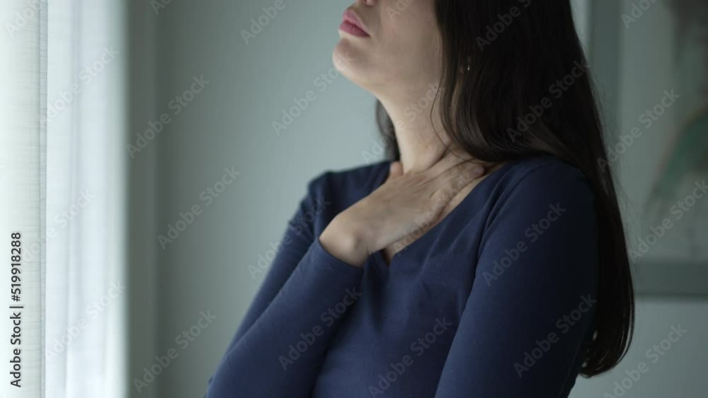 Worried woman standing by window touching her neck and chest seeking comfort. Thoughtful preoccupied person Worried woman standing by window touching her neck and chest seeking comfort. Thoughtful preoccupied person
