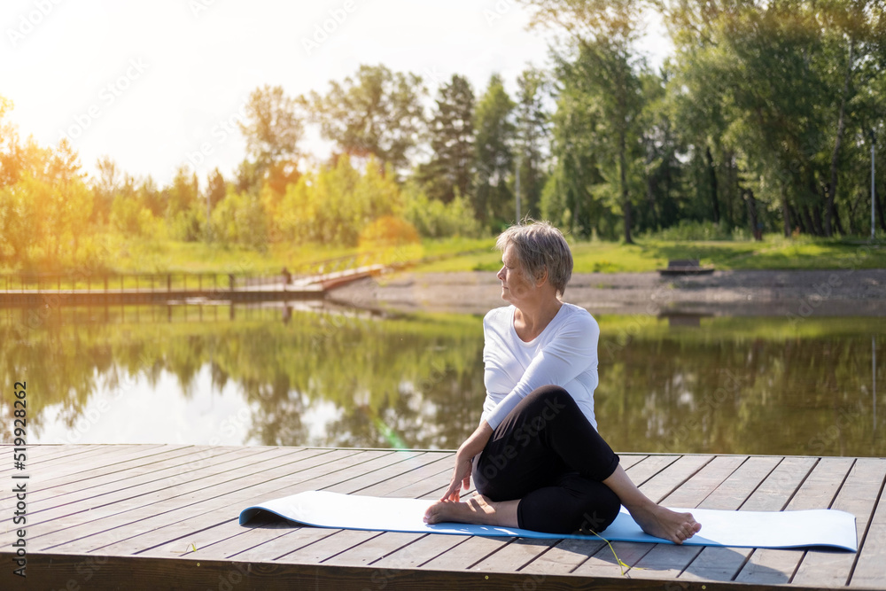 An elderly Caucasian, gray-haired woman does stretching exercises in the park on a yoga mat. Outdoor sports in summer at dawn. The concept of the health of the elderly.