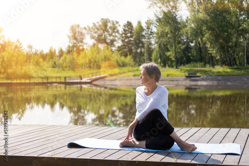 An elderly Caucasian, gray-haired woman does stretching exercises in the park on a yoga mat. Outdoor sports in summer at dawn. The concept of the health of the elderly.