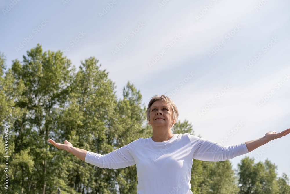 An elderly Caucasian woman stands with her hands raised against the background of green trees and the sky in the park. the concept of success, self-acceptance. An elderly Caucasian woman stands with her hands raised against the background of green trees and the sky in the park. the concept of success, self-acceptance.