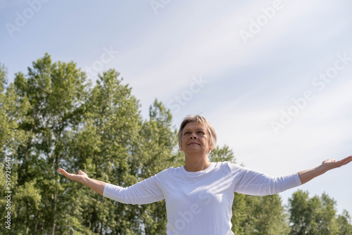 An elderly Caucasian woman stands with her hands raised against the background of green trees and the sky in the park. the concept of success, self-acceptance.