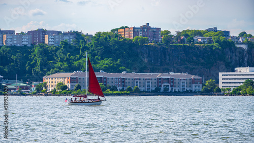 A small sailboat with a red sail, travels north up the Henry Hudson River