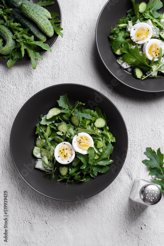 Green summer salad with arugula, cucumbers and egg on black plate. Top view food with Sunny light.