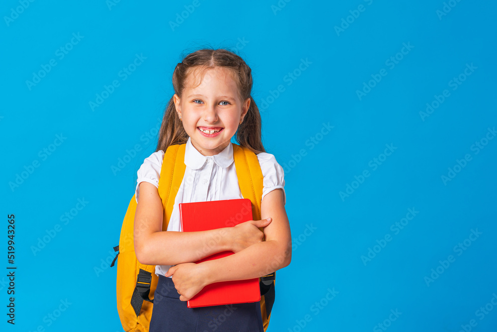 portrait of a funny smiling schoolgirl with a backpack and a book in her hands, posing on a blue background. Back to school. a positive little girl is happy to start classes. Advertising portrait of a funny smiling schoolgirl with a backpack and a book in her hands, posing on a blue background. Back to school. a positive little girl is happy to start classes. Advertising