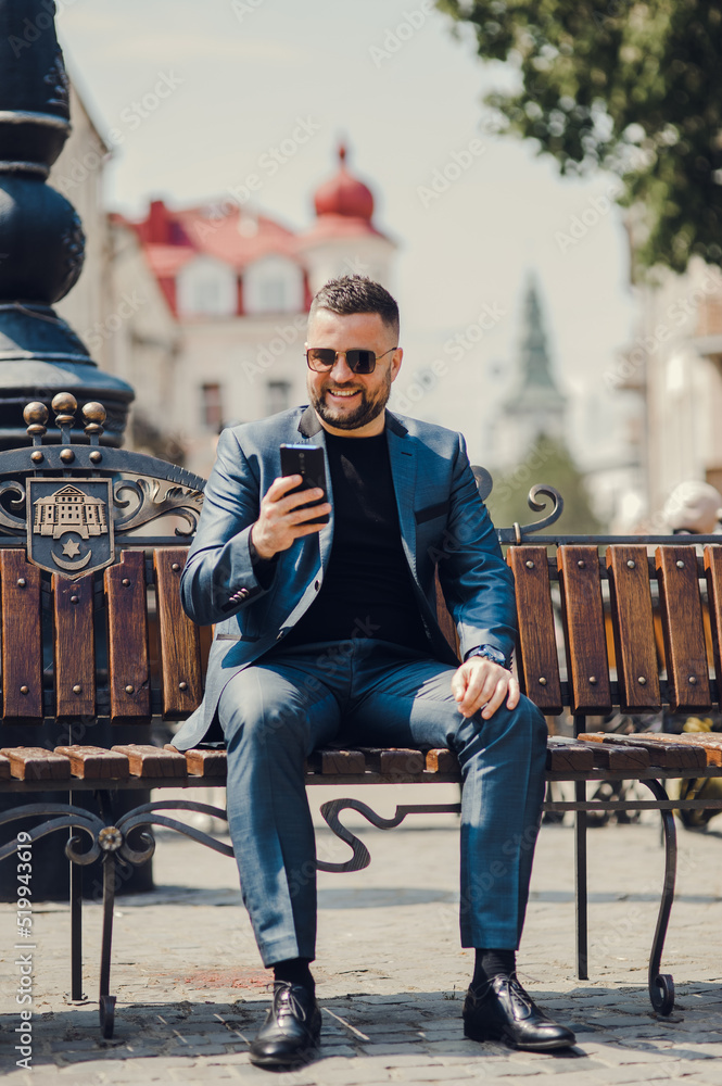 Obraz premium A man in a stylish suit is sitting on a bench, talking to a friend on the phone. Having fun.A handsome man talk emotionally on messenger.
