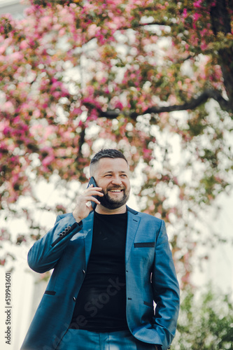 The man waits for a meeting near office. The businessman speaks by phone.Stylish man in a blue suit is standing in the park. Business style of emotion. 