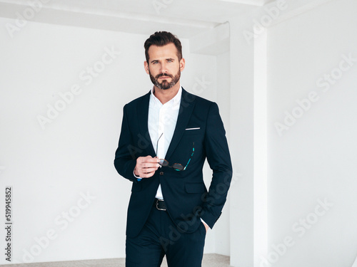 Portrait of handsome confident stylish hipster lambersexual model. Sexy modern man dressed in black elegant suit. Fashion male posing in studio in white interior in sunglasses. Isolated