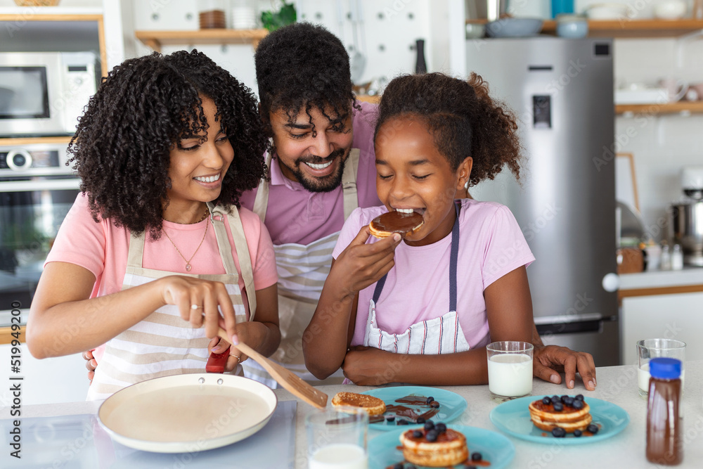 © Graphicroyalty - Family At Home Eating Breakfast In Kitchen Together, Happy African American family standing around stove, daughter eating pancakes