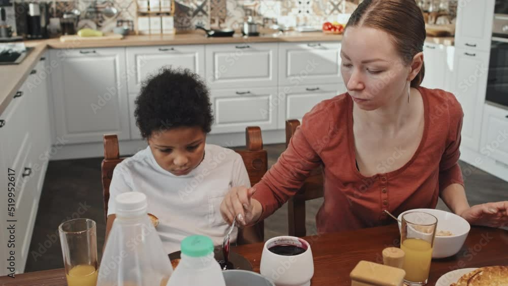 Tilting up slow motion of young Caucasian woman putting jam for pancakes on plate of little African American son, sitting at dining table Tilting up slow motion of young Caucasian woman putting jam for pancakes on plate of little African American son, sitting at dining table
