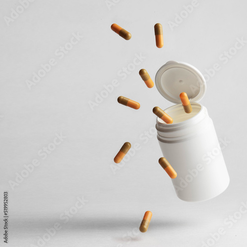 An orange-beige colored pill capsule falling out of a white plastic pill bottle on a light background.