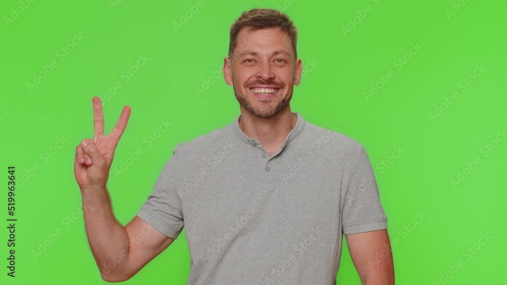 Bearded happy young man showing victory sign, hoping for success and win, doing peace gesture, smiling with kind optimistic expression. Adult guy isolated on green chroma key studio background indoors Bearded happy young man showing victory sign, hoping for success and win, doing peace gesture, smiling with kind optimistic expression. Adult guy isolated on green chroma key studio background indoors