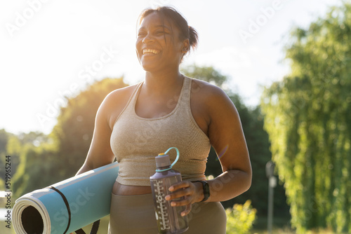 Young African American woman walking with exercise mat and bottle of water through the park in a summer day photo