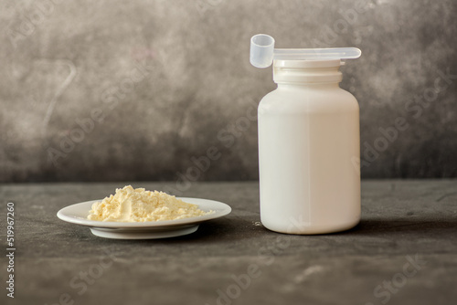 Heap of bovine colostrum powder on plate and white plastic container with measuring scoop on dark background. Colostrum health benefits concept photo