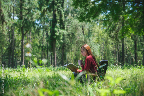 Portrait of a beautiful young asian woman reading a book and drinking coffee while sitting on a camping chair in the park