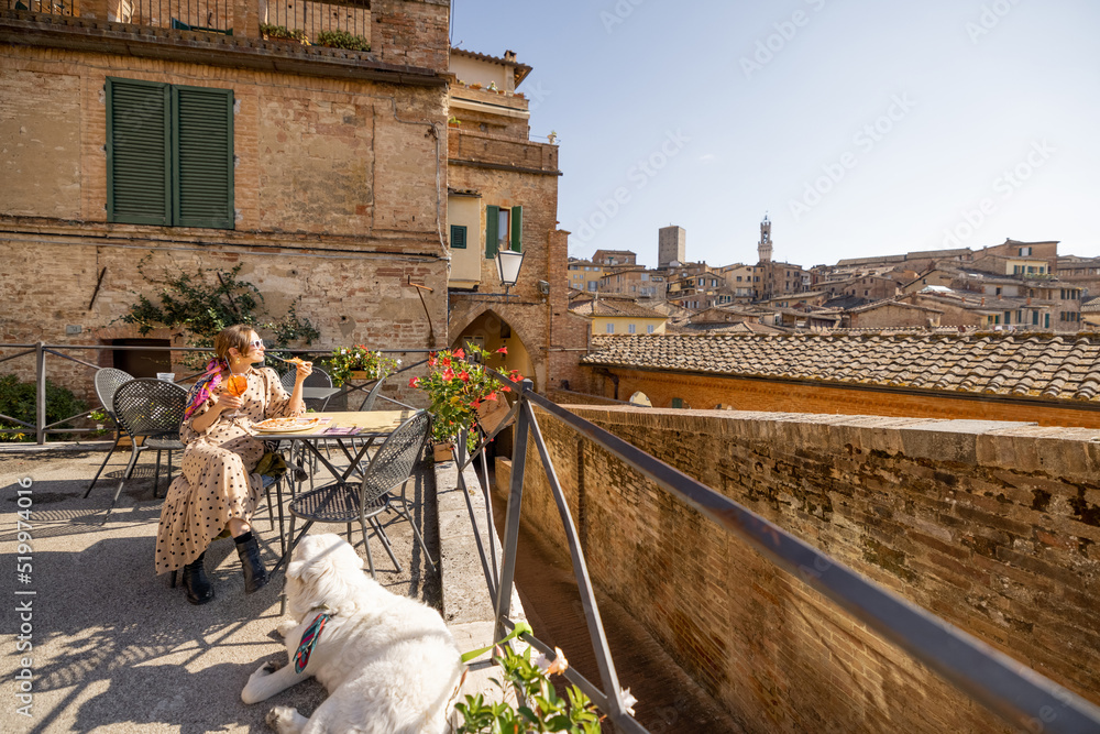Fototapeta premium Young woman having lunch with pizza and wine, sitting with dog at outdoor restaurant with beautiful view on the old town of Siena. Concept of italian cuisine and travel