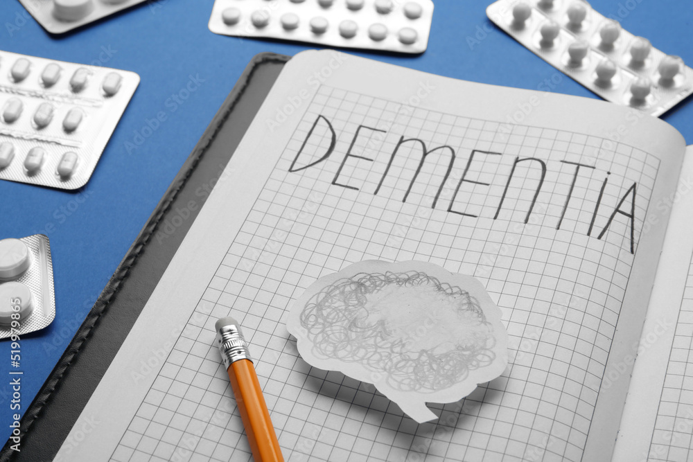 Notebook with word Dementia, brain cutout, pencil and pills on blue background, closeup Notebook with word Dementia, brain cutout, pencil and pills on blue background, closeup