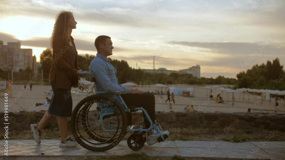 Young woman is driving a young man in a wheelchair along the beach.Disabled guy in the background of the beach in good spirits rides in a wheelchair along the promenade of the city Young woman is driving a young man in a wheelchair along the beach.Disabled guy in the background of the beach in good spirits rides in a wheelchair along the promenade of the city