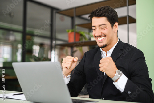 Excited lucky hispanic young businessman sitting at the desk in front of laptop in office and rejoicing with good news, overjoyed male office employee celebrating job offer, clenched fists in triumph
