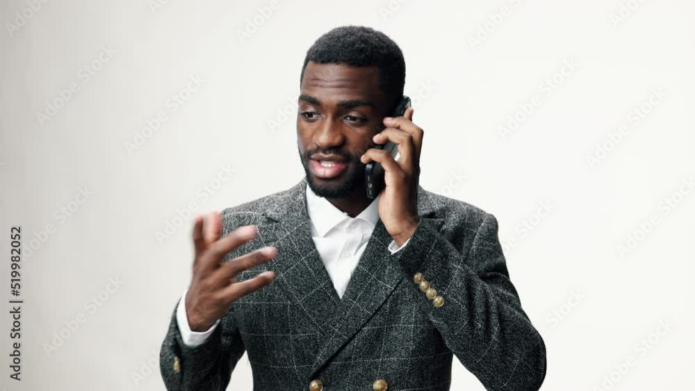 An African-American businessman talking angrily and aggressively on the phone against a gray wall in a business suit and gesturing with his hands An African-American businessman talking angrily and aggressively on the phone against a gray wall in a business suit and gesturing with his hands