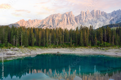 Stunning view of Carezza Lake (Lago di Carezza) with its emerald green waters, beautiful trees and mountains in the distance during a dramatic sunset. © Travel Wild