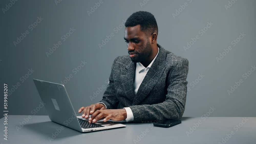 An African American businessman is sitting in an office at his desk with a laptop in a business suit and smile is working on a project An African American businessman is sitting in an office at his desk with a laptop in a business suit and smile is working on a project