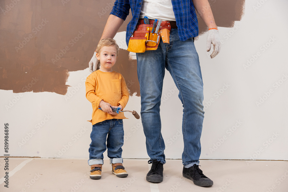 A little blond boy aged 3 years, standing at the feet of his dad. The child holds a paint roller in his hands. His hands are stained with paint. For a child, this is the first experience of repair. A little blond boy aged 3 years, standing at the feet of his dad. The child holds a paint roller in his hands. His hands are stained with paint. For a child, this is the first experience of repair.