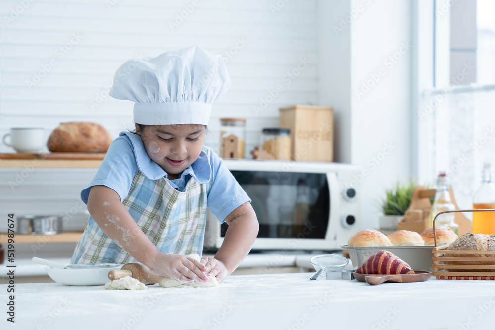 Asian little cute kid girl wear chef hat and apron with flour mess up on face and arms kneading dough bread by her hands in kitchen at home or cooking class at school. Child education concept Asian little cute kid girl wear chef hat and apron with flour mess up on face and arms kneading dough bread by her hands in kitchen at home or cooking class at school. Child education concept