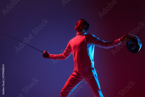 Portrait of young tall male fencer in fencing costume and mask holding smallsword isolated on purple background in neon. Sport, emotions, energy, skills