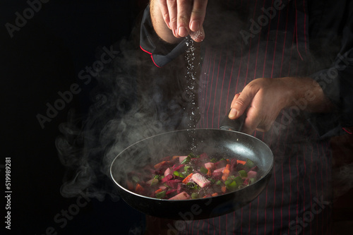 Fototapeta Naklejka Na Ścianę i Meble -  An experienced chef salts vegetables in a hot frying pan. Menu concept for hotel on black background