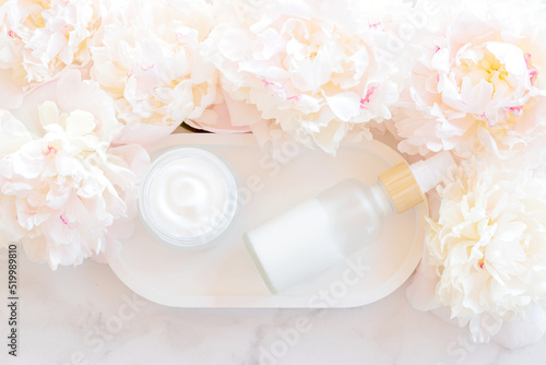 Face and eye moisturizer cream and serum in a glass bottle and jar and peony flowers on light marble background. Concept eco organic natural cosmetic products for skincare