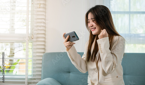 Excited woman use mobile phone raising fist up she celebrating victory win online or received good news on sofa living room at home, Happy Asian female funny on smartphone feeling surprise excited