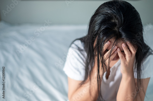Asian woman in white shirt sitting in bed holding her head expressing stress, disappointment, depression, migraines, health issues, health care and stress management concept.