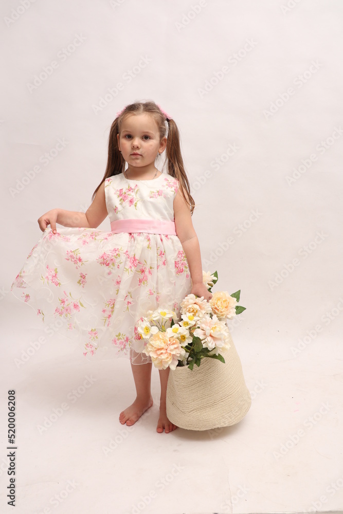 A child on a white background in an elegant light dress. Little girl with a basket of flowers. The girl is waiting to be photographed. Posing. Pampering. A child on a white background in an elegant light dress. Little girl with a basket of flowers. The girl is waiting to be photographed. Posing. Pampering.