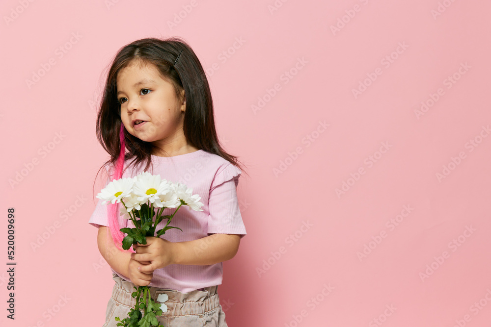 a little beautiful preschool girl stands on a pink background in a pink T-shirt smiling cheerfully holding a bouquet of white daisies in her hands standing sideways to the camera a little beautiful preschool girl stands on a pink background in a pink T-shirt smiling cheerfully holding a bouquet of white daisies in her hands standing sideways to the camera