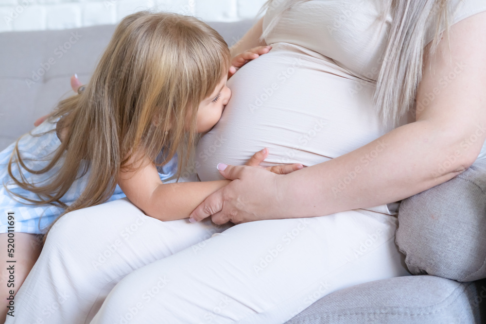 Pregnant woman sitting on bed with little baby daughter stroking and kissing tummy Pregnant woman sitting on bed with little baby daughter stroking and kissing tummy