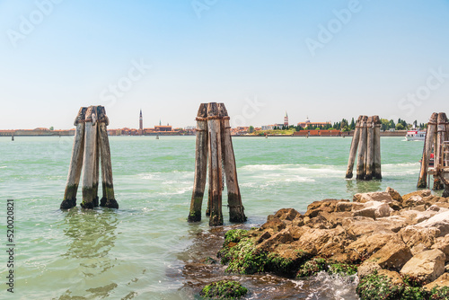 Another angle on the coast of the city of Venice from the opposite side of the canal with a rocky shore, near the wooden poles of the way signs for boats