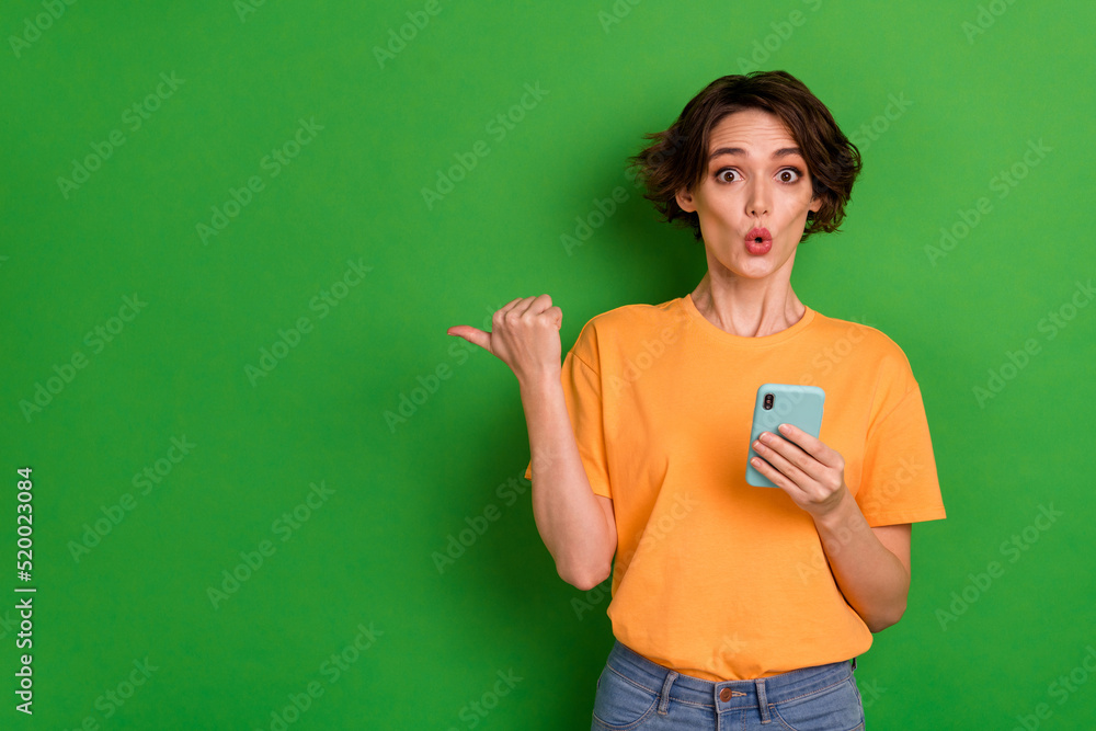Photo of shiny shocked lady wear orange outfit pointing thumb empty space reading modern gadget isolated green color background Photo of shiny shocked lady wear orange outfit pointing thumb empty space reading modern gadget isolated green color background