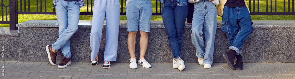 Legs of friends. Legs of six young people in stylish denim clothes standing in row on city street. Horizontal image of male and female legs in different denim pants and shorts. Panoramic banner.