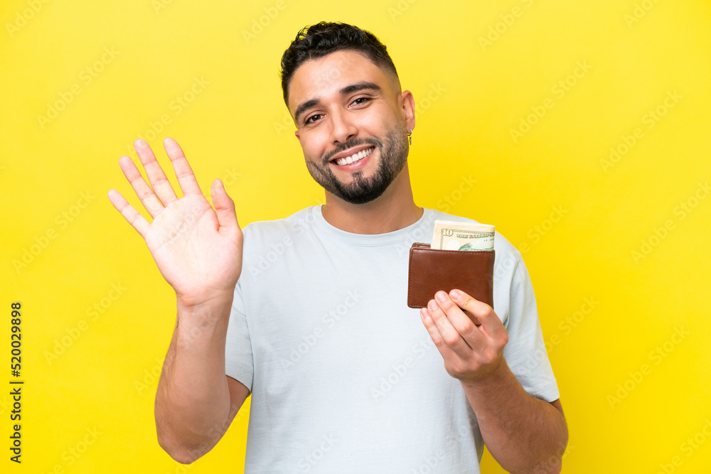 Young Arab man holding a wallet isolated on yellow background saluting with hand with happy expression Young Arab man holding a wallet isolated on yellow background saluting with hand with happy expression