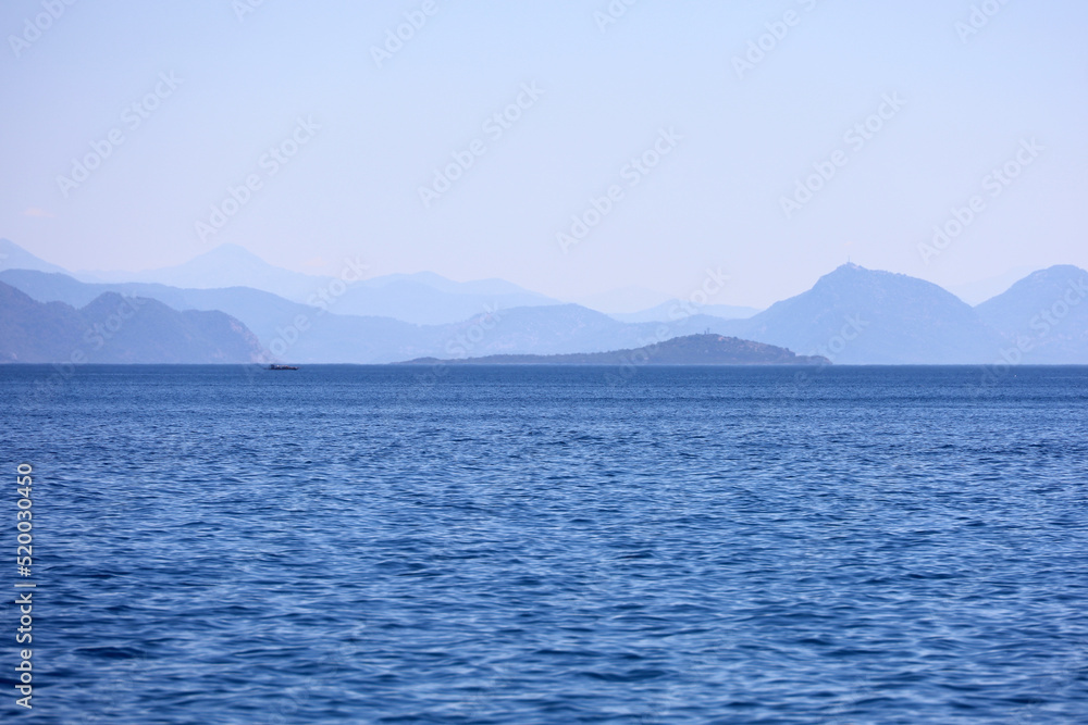 Picturesque view to blue sea and mountain island on horizon in mist. Calm water surface, background for traveling and vacation
