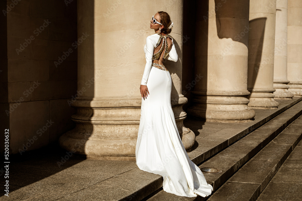 Fototapeta premium Elegant luxury fashion. Glamour, stylish elegant woman in long gown white dress is posing outdoor. Female model in amazing long dress in the park. Outdoor shoot. Vogue. Couture.