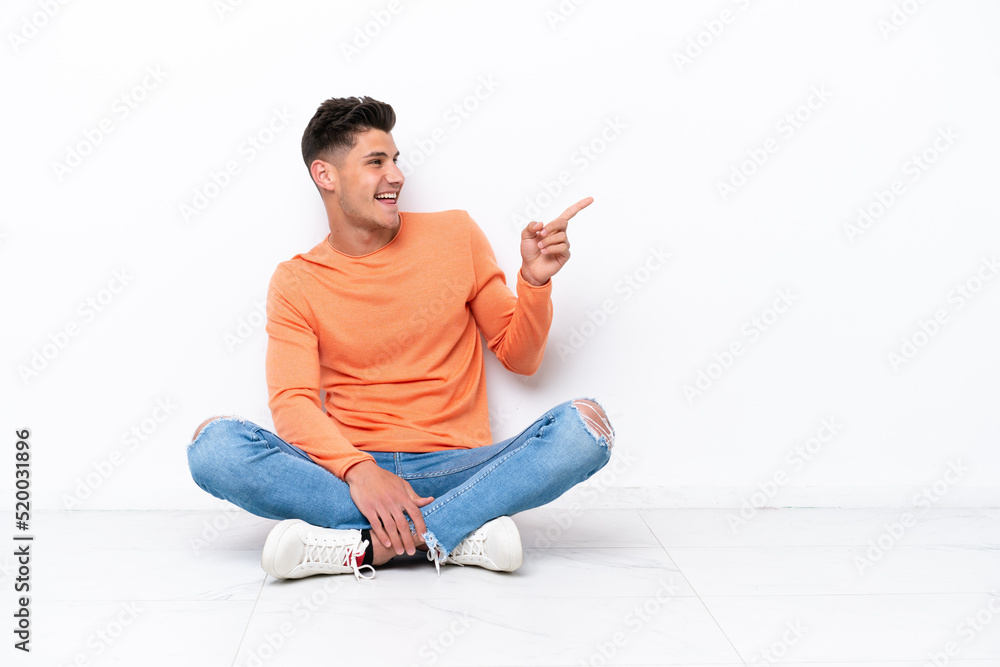 Young man sitting on the floor isolated on white background pointing finger to the side and presenting a product Young man sitting on the floor isolated on white background pointing finger to the side and presenting a product