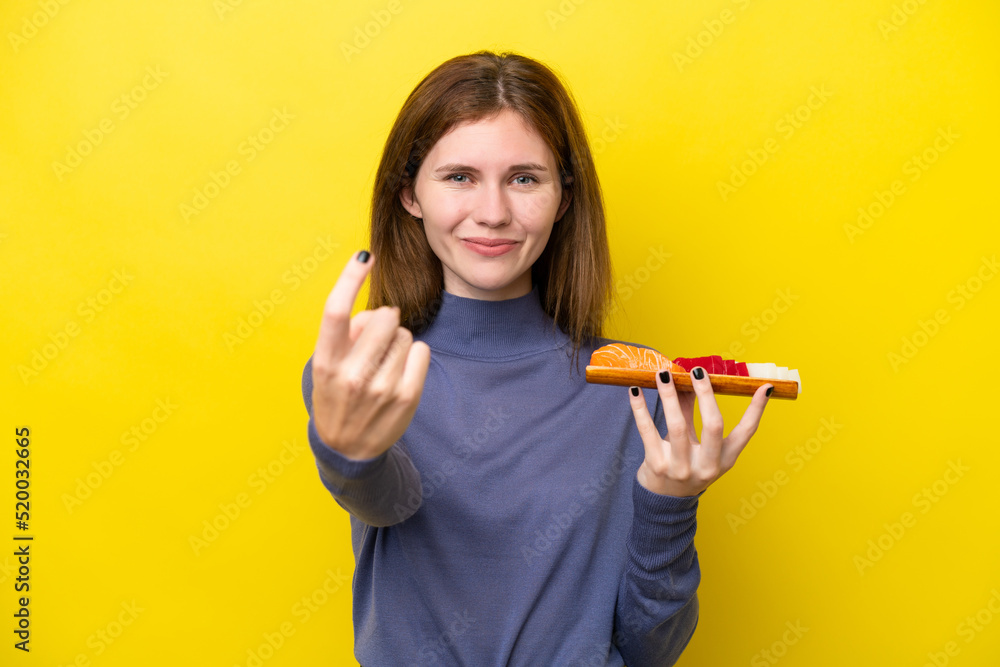 Young English woman holding sashimi isolated on yellow background doing coming gesture Young English woman holding sashimi isolated on yellow background doing coming gesture