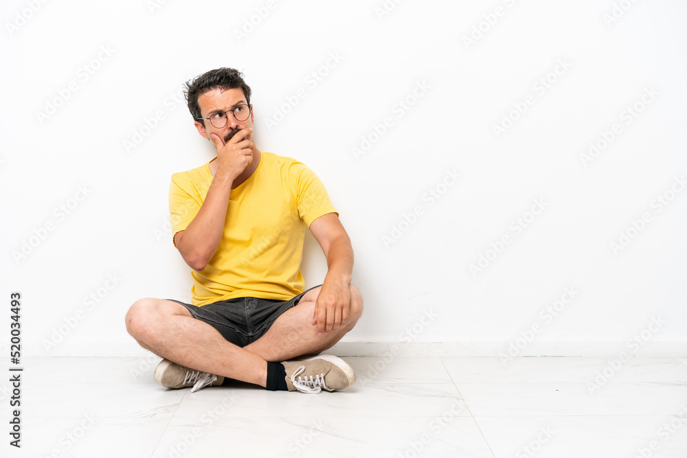 Young caucasian man sitting on the floor isolated on white background having doubts and with confuse face expression Young caucasian man sitting on the floor isolated on white background having doubts and with confuse face expression