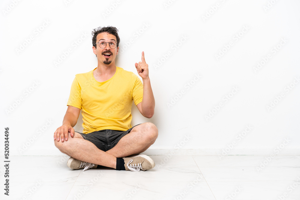 Young caucasian man sitting on the floor isolated on white background pointing up and surprised Young caucasian man sitting on the floor isolated on white background pointing up and surprised