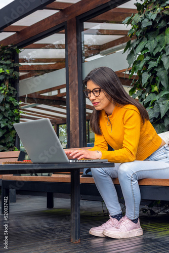 Young hispanic female student with indigenous features wearing a yellow sweater sitting on a bench outside using her laptop